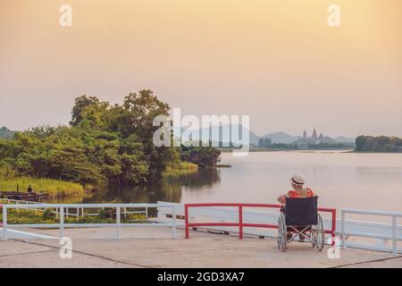 Back view of Asian elderly female patient wearing surgical mask and sitting on wheelchair with feeling lonely and depressed at riverside in the evenin Stock Photo