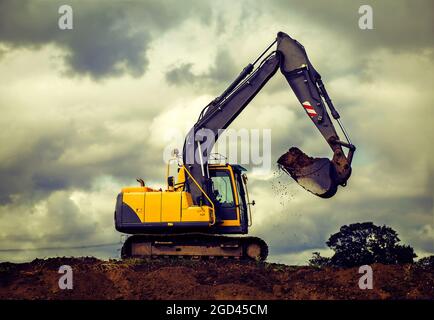 Yellow digger on hill lifting dirt against a dramatic cloudy sky Stock Photo