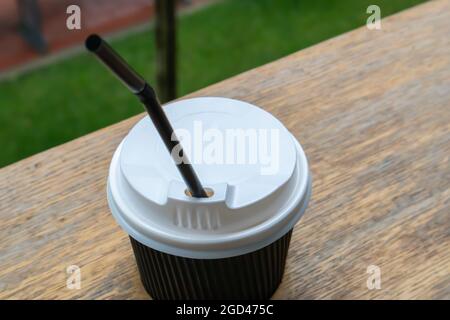 One black paper coffee cup with lid outdoors in summer in sunny weather on a wooden table of a cafe, coffee shop or restaurant Stock Photo