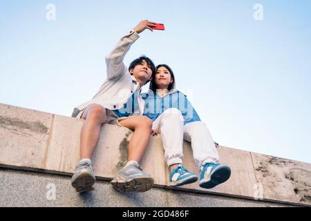 Happy young asian couple sitting in the city posing and taking selfie with their phone. Stock Photo