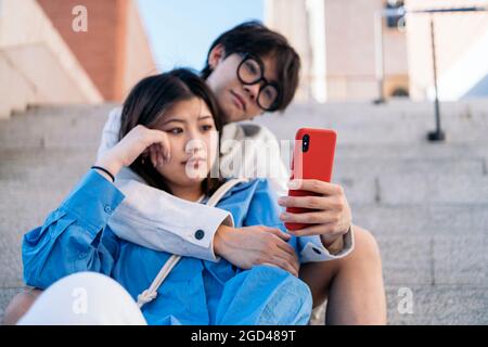 Happy asian couple sitting in the street together and looking at their smart-phone. Stock Photo