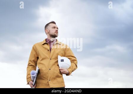 Supervising on site concept. Serious cute caucasian bearded construction contractor, supervisor, foreman or architect with blueprints, laptop and helmet in hands at construction site in orange jacket Stock Photo