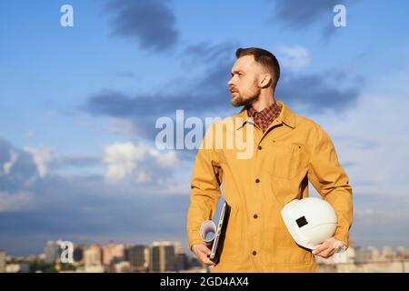 Construction foreman, supervisor, engineer or building contractor. Male in orange jacket standing and holding helmet, laptop and blueprints on construction site with blue sky and big city background Stock Photo