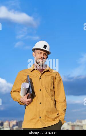 Man in white helmet and orange jacket standing on construction site. Cute caucasian bearded foreman, manager, architect or building contractor at sunny day with blue cloudy sky and urban big city view Stock Photo