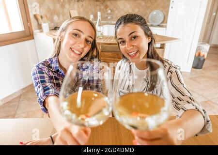 POV portrait of two girls looking at camera holding champagne glasses in selective focus. Students celebrating online with alcohol using video confere Stock Photo