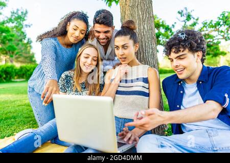 Young hipster friends group sitting on grass in city park making surprise faces looking laptop screen. Modern fun concept with millenials on new trend Stock Photo