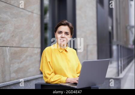 Female young entrepreneur working outside office building in big city. Positive skilled successful business lady staying outdoor at the street with laptop, looking at camera. High quality image Stock Photo
