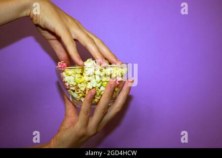 Multi-colored brightly colorful popcorn in a transparent bowl. A woman's hand reaches out, takes sweet popcorn from a glass bowl on a purple backgroun Stock Photo