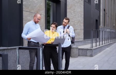Designers or architect team looking at project blueprint and laptop as presentation outdoors. Business persons, men and woman in casual clothes standing outside, brainstorming project Stock Photo