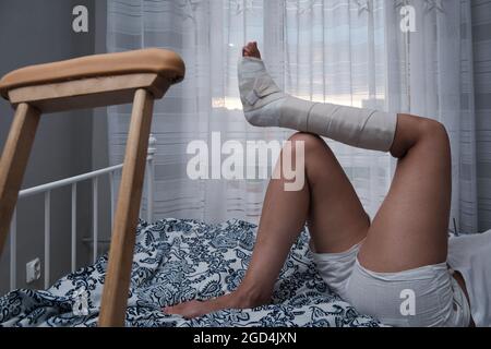 Woman in a plaster cast lies on the bed at home, crutches lie nearby. High quality photo Stock Photo