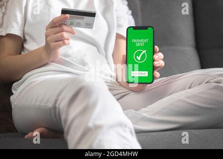 Money transfer concept, woman with credit card sititng on sofa at the home and using online mobile banking, smartphone with payment sent inscription Stock Photo