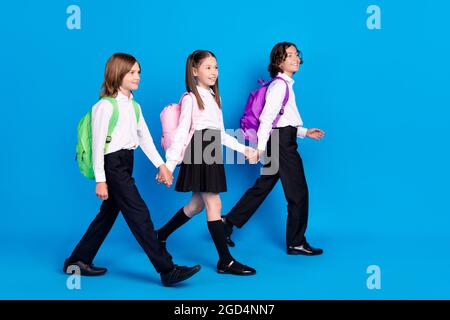 Photo of funny charming schoolchildren wear uniform rucksacks holding arms walking smiling isolated blue color background Stock Photo