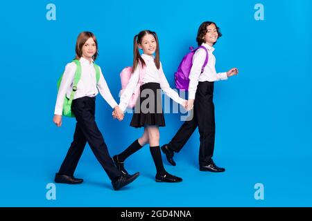 Photo of excited sweet schoolkids formalwear backpacks smiling walking holding arms isolated blue color background Stock Photo