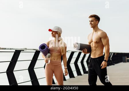 Young fitness couple go to workout, man and woman with fitness mats in hands, getting ready to workout outdoors on the pier, sport and healthy lifesty Stock Photo