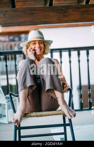 Mature caucasian blonde woman in her 50s talking on a mobile phone smiling and wearing a hat and sitting in a relaxed pose. Lifestyle concept. Stock Photo