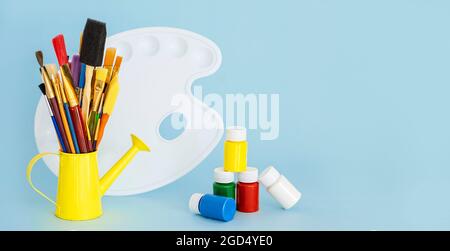 Painting Brushes in yellow mini watering can, variety of paint and artist's white palette on blue background, with copy space Stock Photo