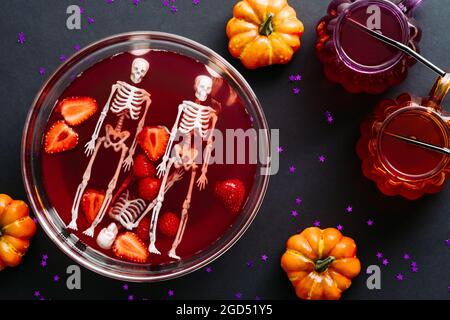 Happy halloween holiday concept. Bowl with blood, strawberries, skeletons and cocktails on dark background. Halloween party flat lay composition. Stock Photo