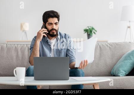 Irritated arab businessman working from home, sitting on couch with laptop, holding documents and having phone call, fighting with assistant or collea Stock Photo