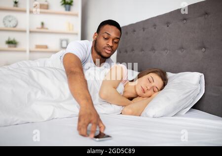 Jealousy, infidelity, relationship problems. Suspicious black husband checking sleeping wife's mobile phone in bed at home. Controlling Afro boyfriend Stock Photo