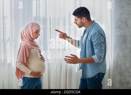 Relationship problems. Aggressive arab man arguing with his upset pregnant muslim wife at home, standing near window, side view. Stressed expectant la Stock Photo