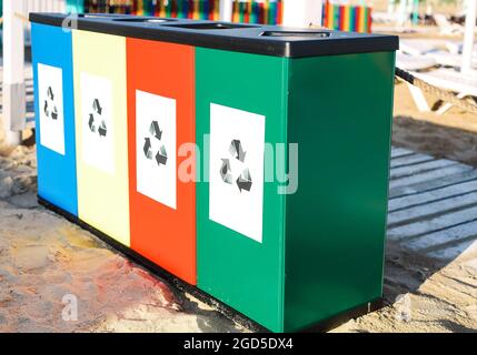 Close-up photo of four different color recycling containers with logos standing on summer beach. Blue, yellow, red and green waste bins for separating Stock Photo