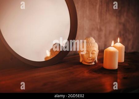Round mirror on wooden shelf with two burning candles and small porcelain Buddha head reflected in corner of mirrored surface over concrete wall backg Stock Photo