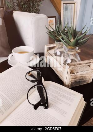 Eyeglasses on open book lying on round coffee table decorated with aloe plants on wooden stand next to cup of tea on saucer, living room interior back Stock Photo