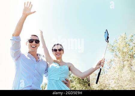 Young excited joyful couple, man and woman dressed festively making selfie with smartphone while sitting on car roof, screaming and smiling from happi Stock Photo