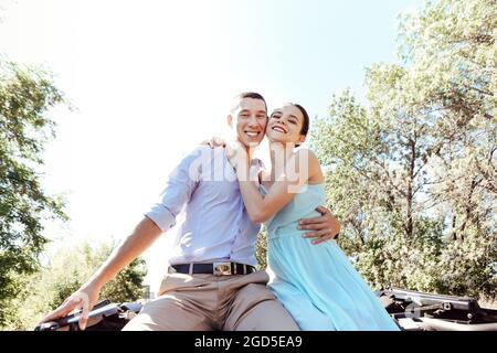 Young excited joyful couple, man and woman dressed festively while sitting on car roof, screaming and smiling from happiness while raising hands up in Stock Photo