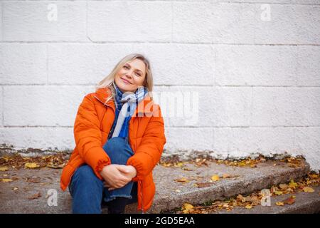 Happy smiling middle aged woman in orange down jacket sitting on concrete stairs outdoors and looking at camera with positive face expression, resting Stock Photo