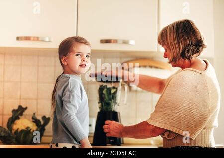 Loving grandmother cooking together with granddaughter at home, using blender to mix vegetables, little girl learning how to use appliances with grand Stock Photo