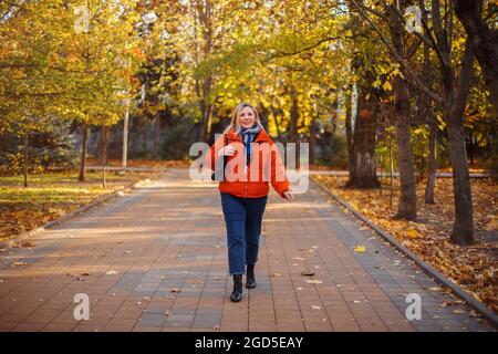 Happy positive middle-aged woman in orange down jacket smiling at camera while walking outdoors on sunny winter day, cheerful mature female enjoying l Stock Photo