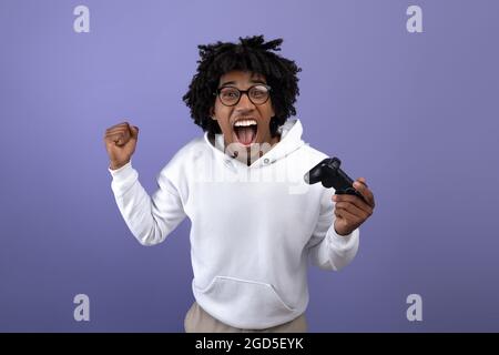 Triumphant black teen guy with joystick shouting in excitement, playing video game, making YES gesture on violet background. Overjoyed teenage gamer w Stock Photo