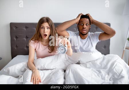 We are late. Shocked interracial couple with alarm clock grabbing their heads in terror, sitting on bed at home. African American guy and his wife fee Stock Photo