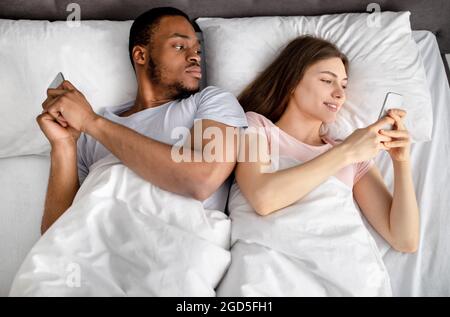 Young interracial couple lying in bed stuck in smartphones, playing online games, browsing social media, top view. Relationship problems, gadget addic Stock Photo