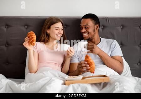 Millennial multiracial couple having breakfast in bed together, drinking coffee with croissants, spending honeymoon at hotel. Multiethnic newlyweds en Stock Photo