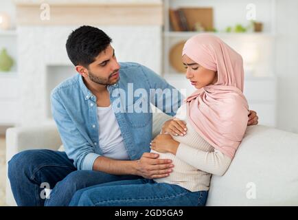 Care in couple during pregnancy. Worried arab husband supporting pregnant wife with prenatal contractions at home. Woman suffering from pain, sitting Stock Photo