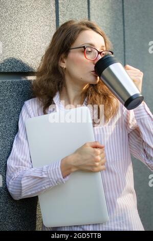 Female portrait of young woman with silver laptop and cup of coffee waiting for a meeting near dark grey wall in the morning, remote job, coffeebreak Stock Photo