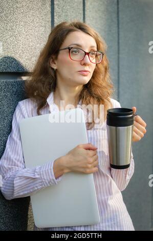 Female portrait of young woman with silver laptop and cup of coffee waiting for a meeting near dark grey wall in the morning, remote job, coffeebreak Stock Photo