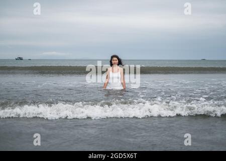 Beautiful young asian woman in white dress standing up to the waist in the sea. Looking at camera with copy space. Romantic photo. Ocean view.  Stock Photo