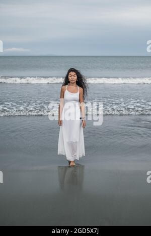Charming young mongol woman in bright grey dress standing on the beach. Black long curly hair. Looking at camera with copy space. Romantic photo. Stock Photo