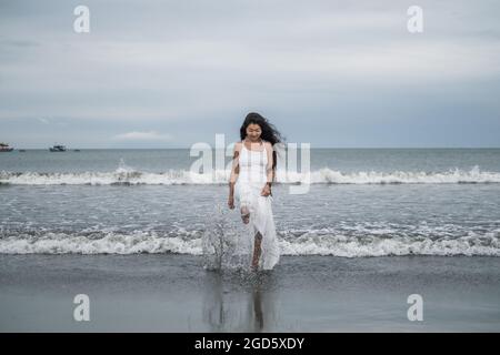 Lovely young asian woman in white dress walking and playing with water on the beach. Kicking the water. Black long curly hair. Romantic photo. Stock Photo