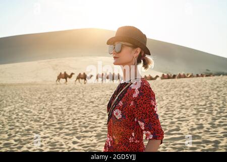 asian woman in red dress looking at view in desert with caravan of camels and huge sand dunes in background Stock Photo