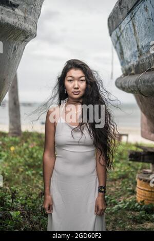 Beautiful young mongol woman in bright grey dress standing between two old wooden ships. Looking at camera with copy space. Romantic photo. Stock Photo