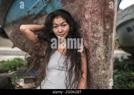 Portrait of beautiful young asian woman in white dress standing near the old ship and holding hair with hand. Natural black curly long hair. Stock Photo