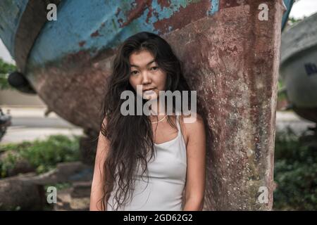 Portrait of beautiful young asian woman in white dress standing near the old ship. Natural black curly long hair. Thinking romantic face.  Stock Photo