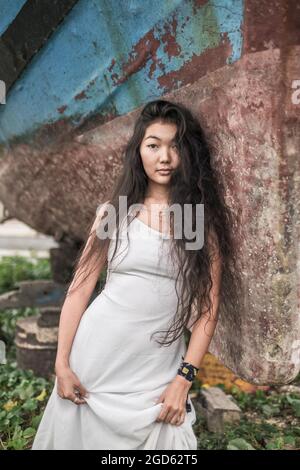 Portrait of charming young mongol woman in white dress standing near the wall. Natural black curly long hair. Looking at camera with copy space. Stock Photo