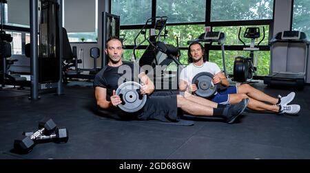 group of friends sitting on a fitness mat in a gym indoor exercising with heavy lifting. two men during hard train practicing together inside a club Stock Photo