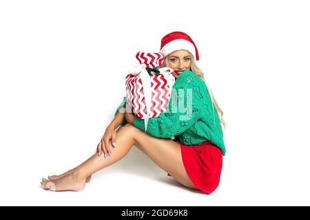 Portrait of sitting barefoot beautiful woman in santa hat, toothy smiley face, white background, copy space, studio shot, weared in green red color, h Stock Photo