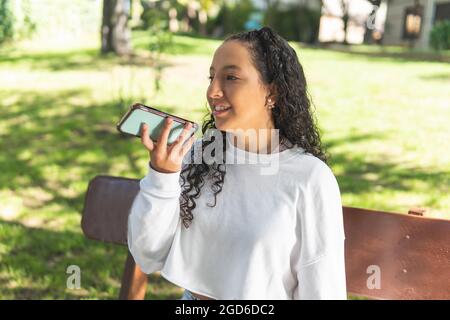 Teenage woman with curly hair sending a voice message with her cell phone sitting on a park bench Stock Photo